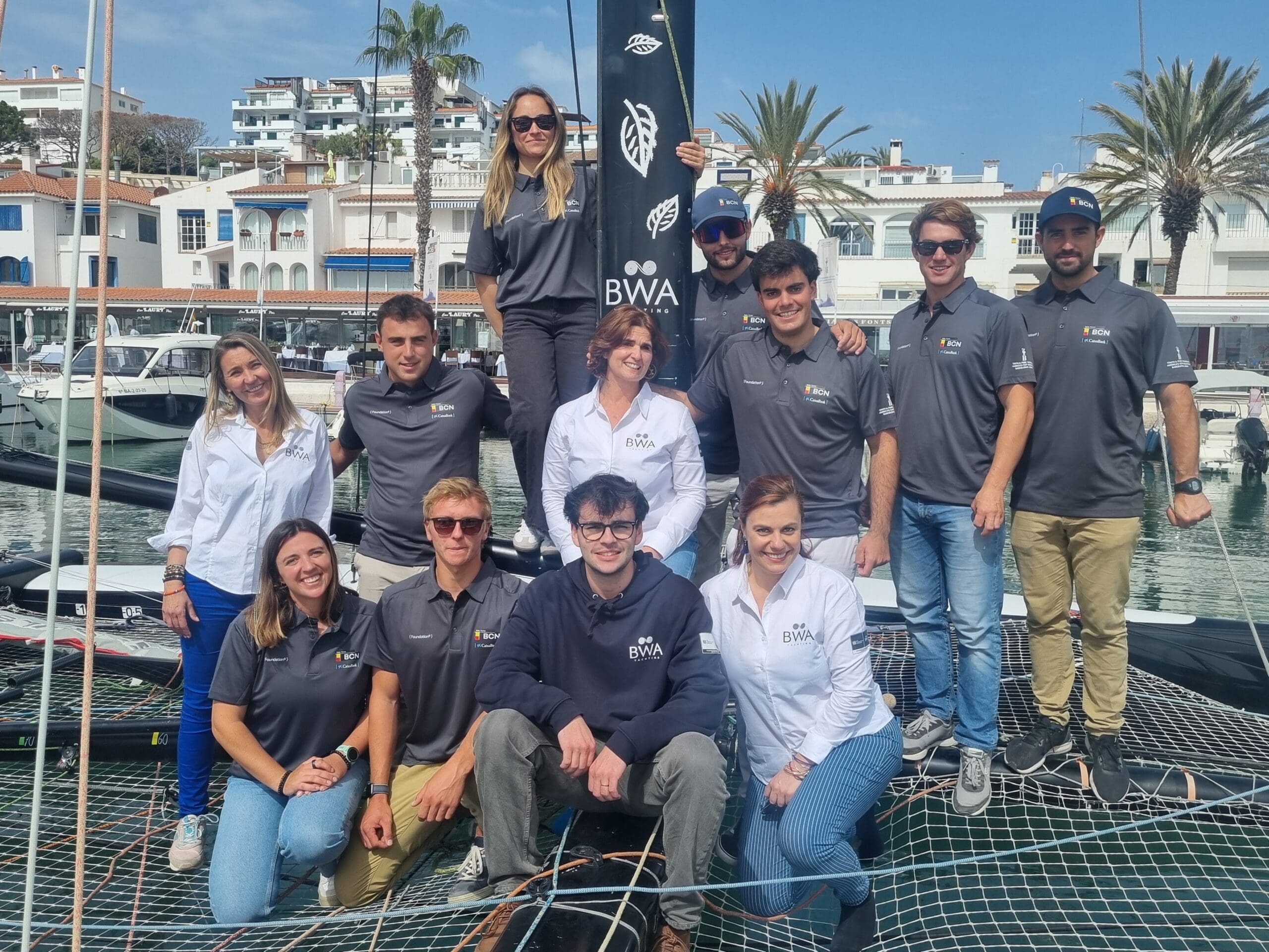 Group photo of 12 men and women taken in the harbour on a superyatch catamaran on the netted area with a BWA Yachting flag in the background.
