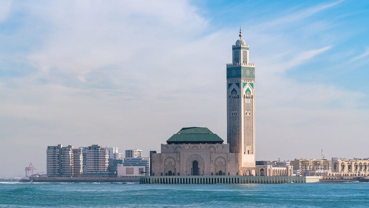 Image of Morocco from the sea of the city. A tall decorative building stands in the middle of the image in white and turquoise. Behind is a backdrop of high rise buildings to the left and smaller buildings to the right. The sky is blue with light clouds.