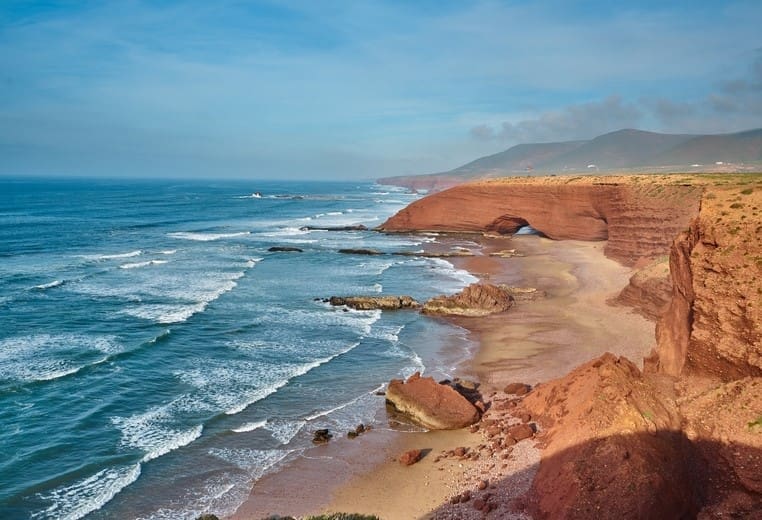 Coastal image of Morocco. The coast on the right is desert like, dusty and orange with a sharp drop down to the beach with sand. Calm blue waters are lapping onto the sandy beach from the right as they break the water is white coming into shore. There are a few large and small rocks on the sand. In the background are large mountains. The sky is blue with a few dark clouds over the mountains.
