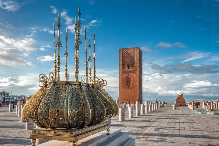 Image of Morocco. Gold metal decorative structure and a brick tower building in the background. The skys are blue with a few clouds with the floor tiled concrete.
