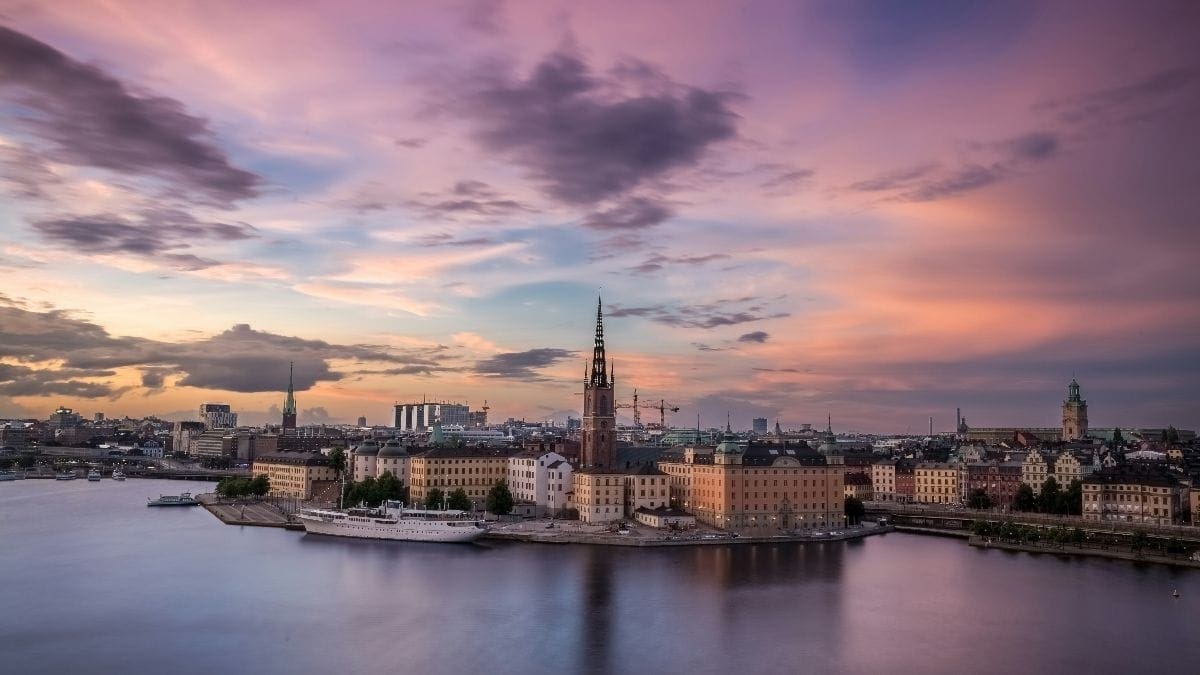 Sunset shot taken from the sea looking at Stockholm, Sweden. The sky is glowing pink with some clouds in the sky. A white luxury superyacht can be seen docked with the rest of the city in the background.