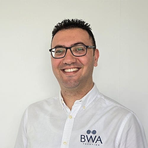 BWA Yachting employee headshot. Man smiling (with his teeth) with dark short brown hair, glasses and a white shirt with the BWA Yachting logo embroidered in blue.