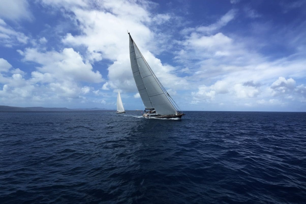 Luxury sailing superyacht in the ocean leaning left in the wind with another luxury sailing super yacht following behind. The ocean is dark and the sky is blue with lots of clouds. The superyacht has plain white sails and a dark blue hull.