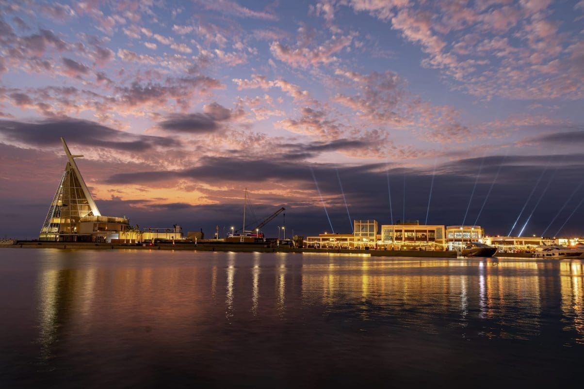 Saudi port at evening with lights projecting into the sky from buildings and port reflected in the water